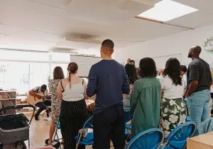 A group of adults of all ages sitting in rows in a school classroom. There is a man playing a guitar and worship lyrics on the screen at the front.